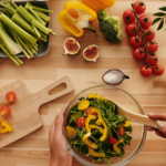 Person preparing a fresh salad with various colorful vegetables on a wooden table.