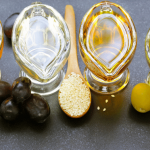 Four glass containers filled with different oils, each accompanied by corresponding seeds or olives on wooden spoons, representing a variety of natural oils.